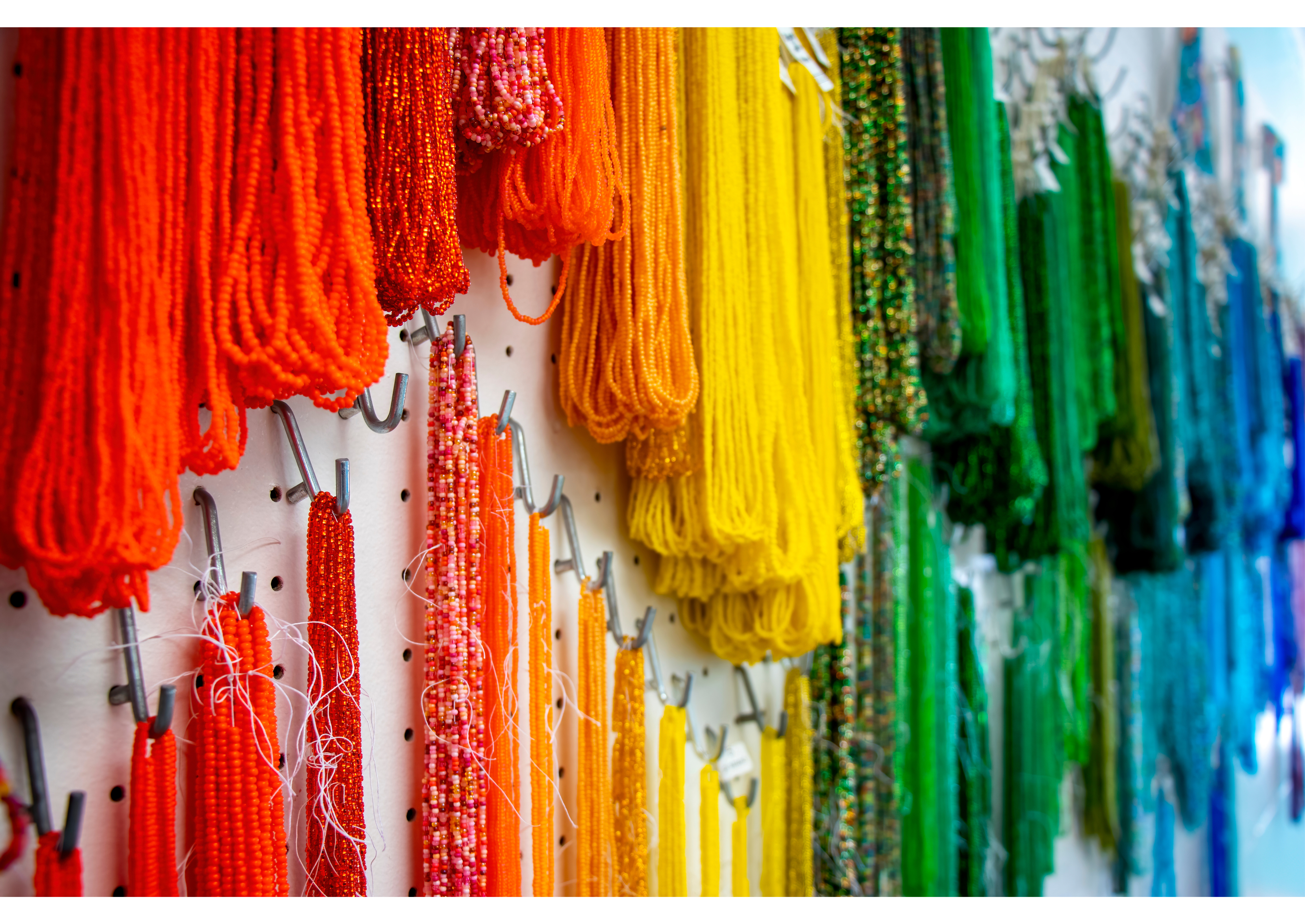 An assortment of hanks of strung beads going from red to orange, to yellow, green, and then blue in a variety of shades hanging on a pegboard.