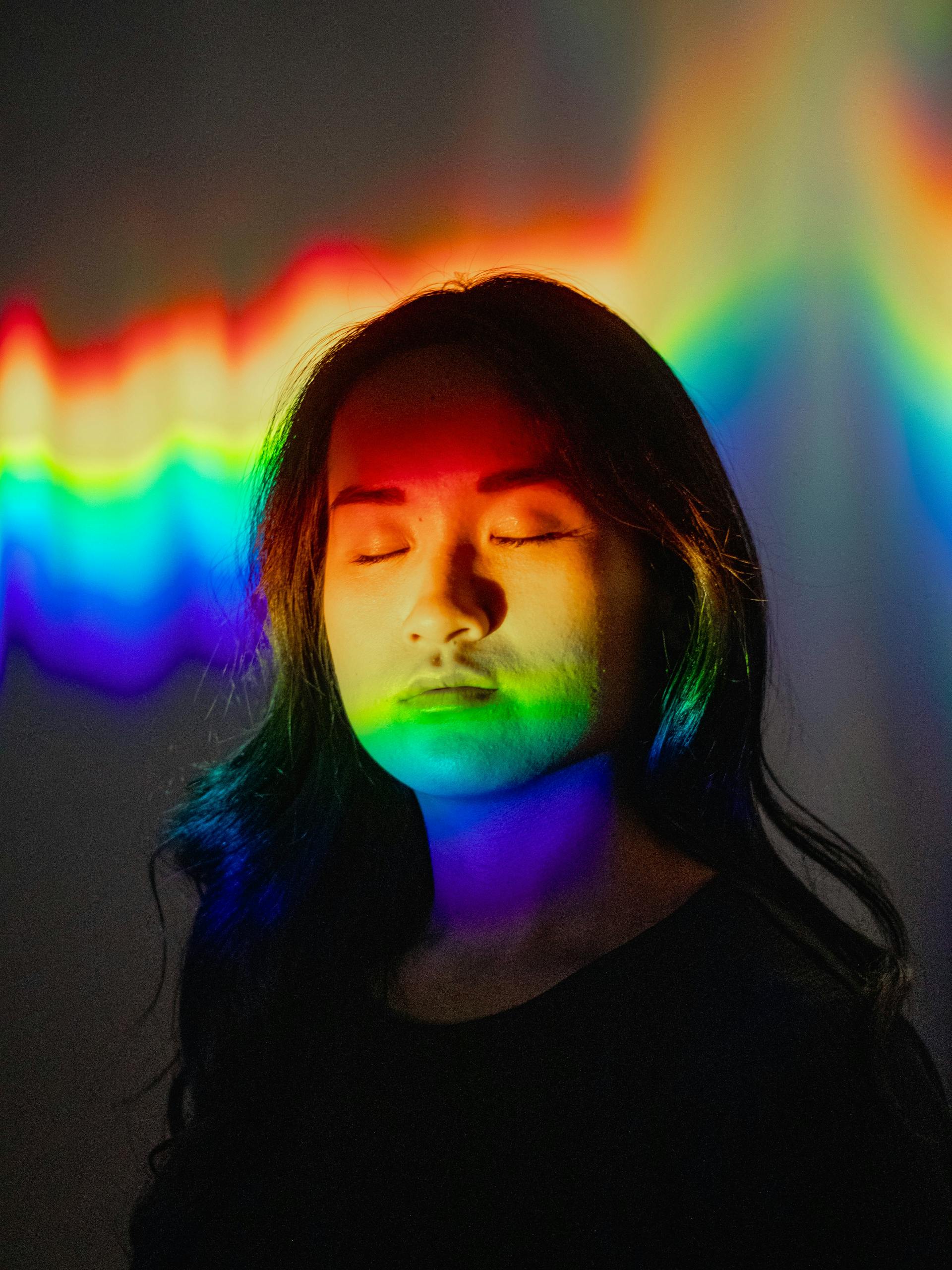 A woman stands in a studio with vibrant rainbow light projections covering her face.